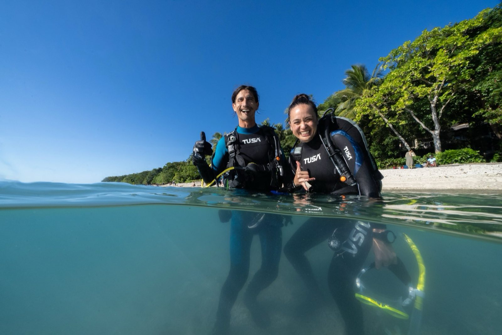 Two divers in the water at the surface beside a tropical shoreline, giving the OK signal.